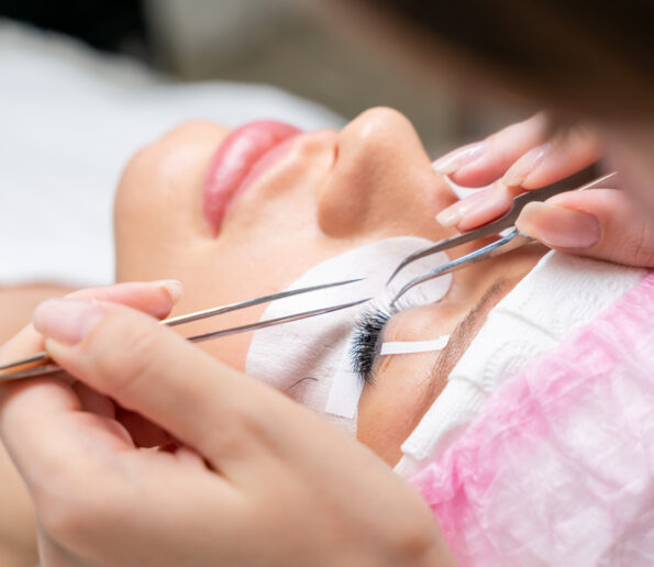 Eyelash extension master with tweezers in his hands performs eyelash correction close-up. Girl's eye with black artificial eyelashes closeup. Eyelash extension procedure in beauty salon. Lashmaker at work. Concept spa lash.